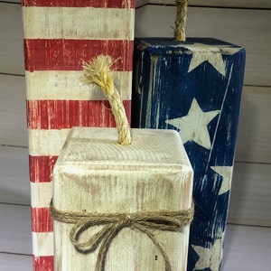 May include: Three rustic wooden firecracker decorations. One is red and white striped, another is blue with white stars, and the third is off-white with a twine bow. Each has a rope fuse.