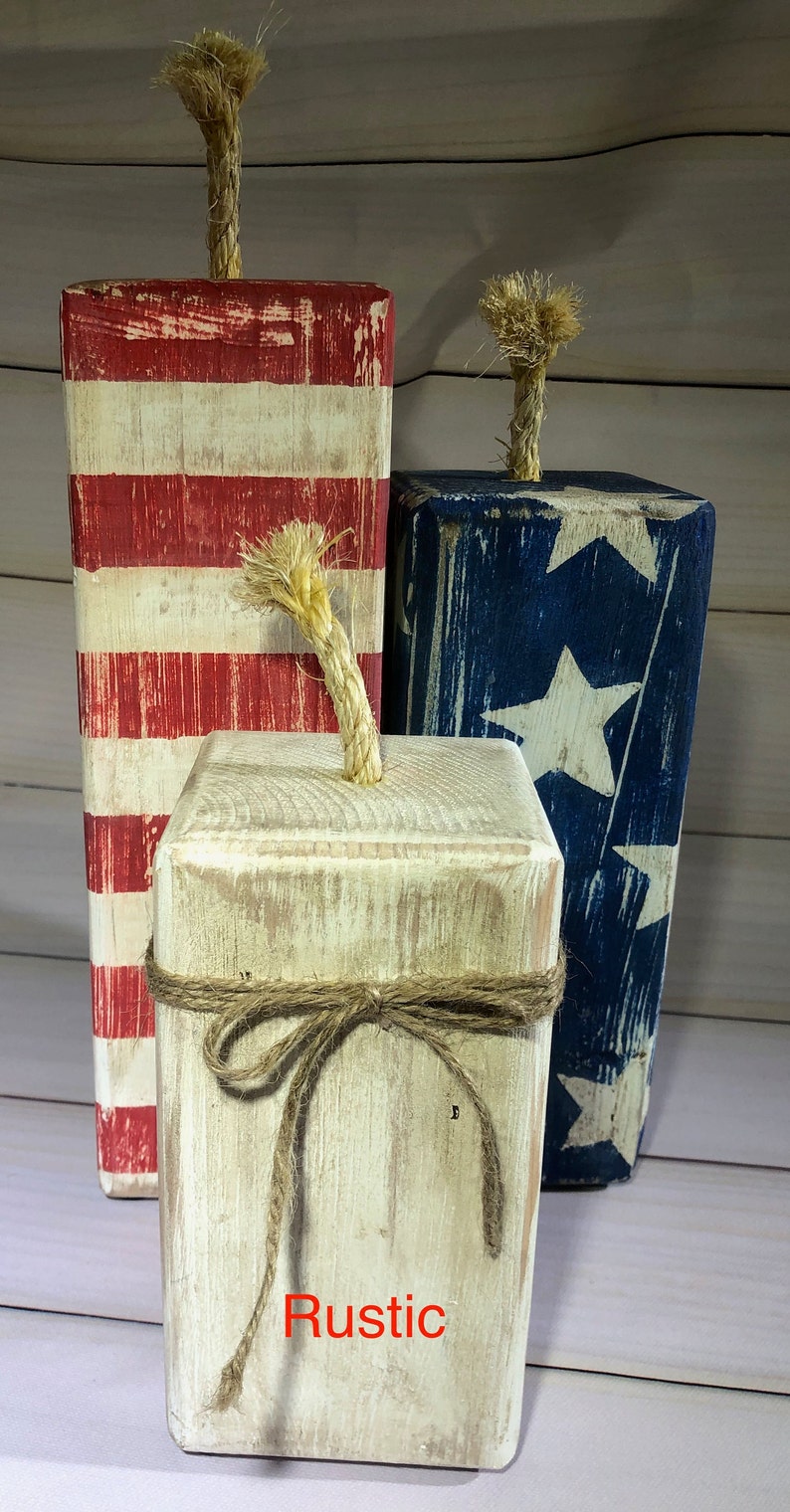 May include: Three rustic wooden firecracker decorations. One is red and white striped, another is blue with white stars, and the third is off-white with a twine bow. Each has a rope fuse.