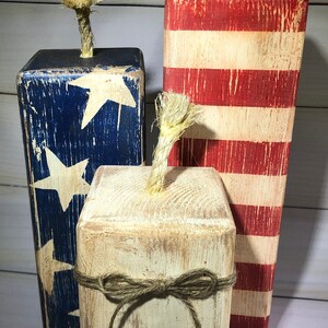 May include: Three wooden firecracker decorations. One is painted with blue and white stars, another with red and white stripes, and the third is off-white with a twine bow. Each has a rope fuse.