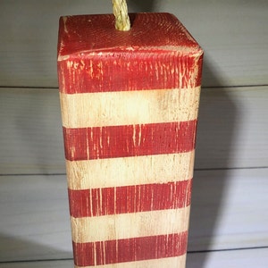 May include: A wooden decorative firecracker with a red and white striped design. The top is painted red and features a natural rope fuse. The rustic, distressed finish gives it a vintage, patriotic aesthetic. Perfect for Independence Day.
