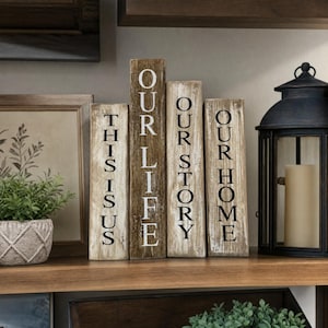 May include: Wooden decorative blocks with the words "THIS IS US", "OUR LIFE", "OUR STORY", and "OUR HOME" printed in black. The blocks are various shades of brown and are arranged on a wooden shelf. A black lantern and a potted plant are also on the shelf.