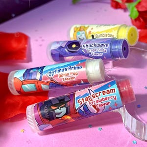 May include: Lip balms featuring Transformers characters. The tubes are decorated with images of Optimus Prime, Bumblebee, Shockwave, and Starscream. Flavors include Bomb Pop, Grape Soda, and Strawberry. The lip balms are on a pink surface with red flower petals.