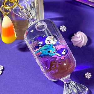 Puede incluir: Amuletos de resina transparentes con forma de caramelo con personajes de dibujos animados en el interior, sobre una superficie morada. Un llavero de estrella dorada con un dije de caramelo de maíz está al lado de un bloque transparente. Elementos decorativos: confeti blanco en forma de estrella y merengues pastel.
