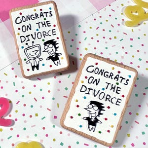 Może przedstawiać: Dwie prostokątne drewniane tabliczki z napisem "Congrats on the Divorce" i postaciami z kreskówek. Tabliczki mają białe tło z kolorowym konfetti i drewnianą ramą. Rozrzucone są dekoracyjne serca.