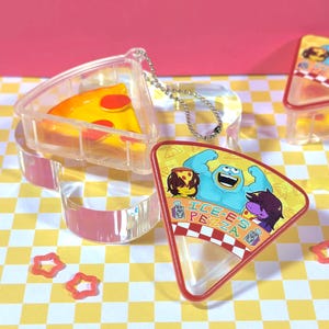 May include: Clear acrylic pizza slice keyrings with a slice of pizza inside. The keyrings have a silver chain. A second keyring with a pizza slice design is visible, with the text "ICE-E PEZZA" and cartoon characters.