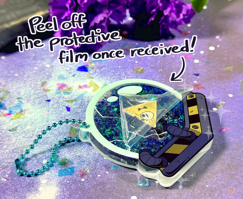 May include: A green and blue glitter filled resin keychain with a yellow triangle with a single eye inside. The keychain is shaped like a glass dome with a black and yellow cartoon character on the bottom. The text "Peel off the protective film once received!" is written on the image.