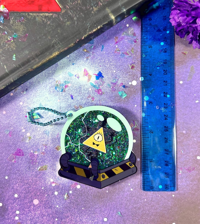 May include: A green and yellow glitter filled glass dome keychain with a cartoon image of a yellow triangle with a single eye and a bow tie inside. The triangle is surrounded by a black and white lightning bolt design.
