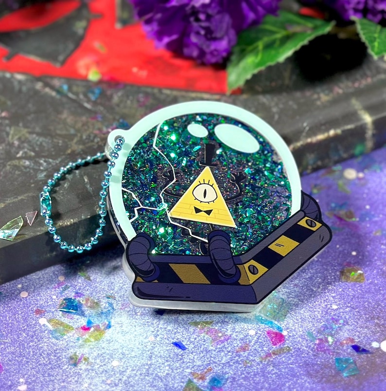 May include: A blue and green glitter filled keychain with a yellow triangle character with one eye and a bow tie inside. The character is inside a clear dome with a black and yellow striped base.