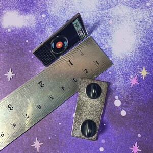 May include: A silver enamel pin featuring a black and white illustration of HAL 9000 from the movie 2001: A Space Odyssey. The pin is on a ruler with the text "HAL 9000" visible. The pin has a black backing with two silver posts.