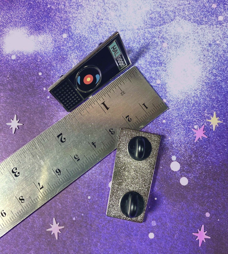 May include: A silver enamel pin featuring a black and white illustration of HAL 9000 from the movie 2001: A Space Odyssey. The pin is on a ruler with the text "HAL 9000" visible. The pin has a black backing with two silver posts.