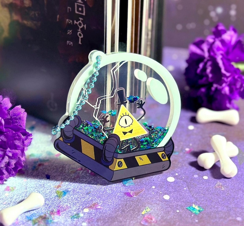 May include: A clear plastic sphere charm with a yellow triangle character inside. The character has one eye and a bow tie. The sphere is filled with glitter and has a silver chain attached.