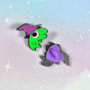May include: Two enamel pins, one green with a purple hat and a large eye, the other a black heart with a purple back.