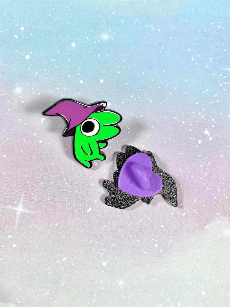 May include: Two enamel pins, one green with a purple hat and a large eye, the other a black heart with a purple back.