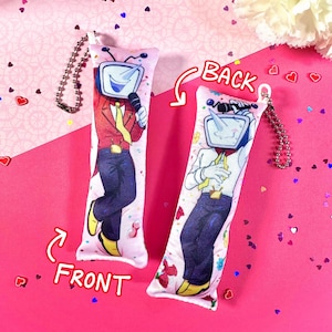 May include: Two rectangular plush keychains with a cartoon TV-headed character. One keychain shows the front of the character in a red jacket, blue pants, and yellow shoes, holding a microphone. The other keychain shows the back of the character in a white shirt and blue pants.