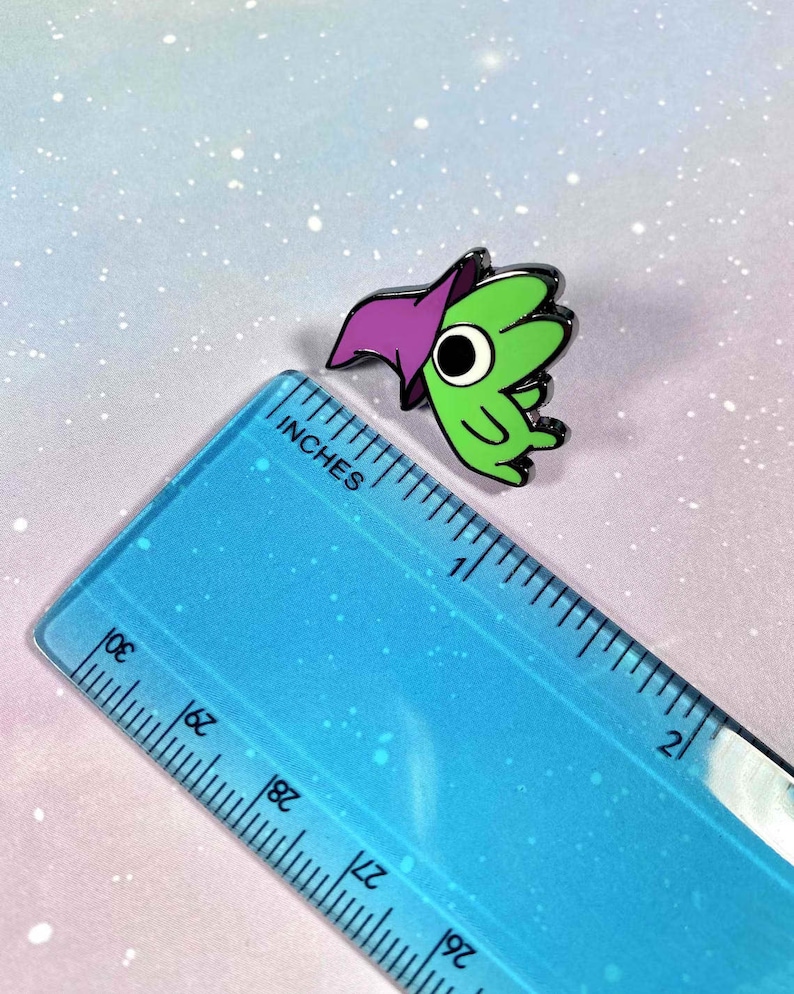 May include: Green and purple enamel pin of a cartoon character wearing a purple hat. The character has one large eye and is about 1 inch tall.