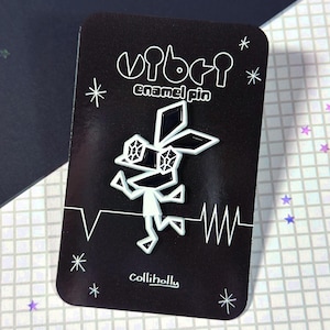 May include: Black and white enamel pin featuring a stylized geometric character with a heart beat graphic. The pin is labeled "vibe" and "colliholly" and says "enamel pin".