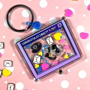 May include: A clear acrylic keychain with a purple frame and a cartoon character inside. The character is a black-haired person with a white shirt and a black tie. The keychain also has a blue and white telephone, hearts, and envelopes inside. The text "CONGRATULATIONS!! <3!" is visible in the frame.