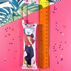 May include: A long, rectangular plush keychain featuring a cartoon character with a TV head, wearing a red jacket, yellow tie, and black pants. The keychain is approximately 10 inches long and has a silver ball chain. The background is pink.