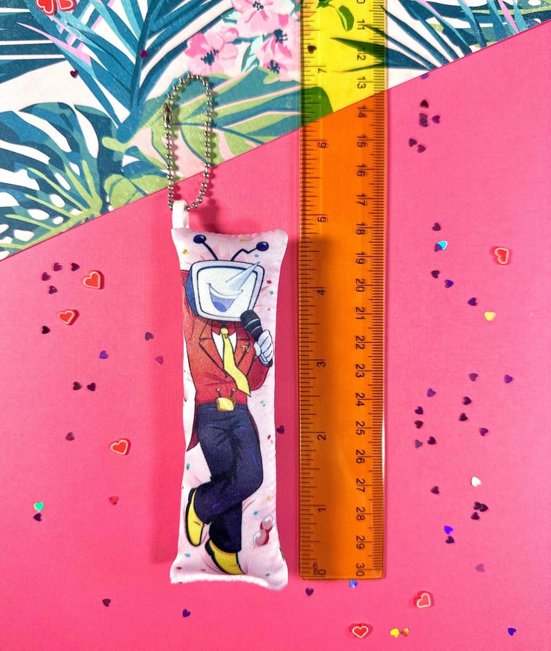 May include: A long, rectangular plush keychain featuring a cartoon character with a TV head, wearing a red jacket, yellow tie, and black pants. The keychain is approximately 10 inches long and has a silver ball chain. The background is pink.