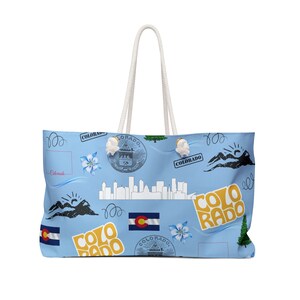 Puede incluir: Una bolsa de lona azul claro con un asa de cuerda blanca. La bolsa presenta un patrón repetido de imágenes con temática de Colorado, incluyendo la bandera del estado, un horizonte, montañas y la palabra "Colorado" en diferentes fuentes.