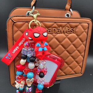May include: A brown handbag with a quilted pattern, paired with a Spider-Man keychain. The keychain features a red Spider-Man figure, a red strap with "Spider-Man" text, and a beaded charm with various colorful beads.