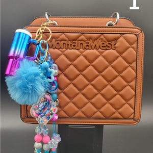 May include: Brown quilted handbag with the text "Montana West". The bag has silver handles and a decorative keychain with a blue pom-pom, a Stitch character charm, and beaded accents. The keychain also includes a blue and purple ombre tumbler.