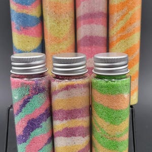 May include: Eight clear glass bottles with silver lids filled with colorful layered sand. The sand is in a variety of colors, including pink, blue, green, yellow, and orange. The bottles are arranged in a row on a black surface.