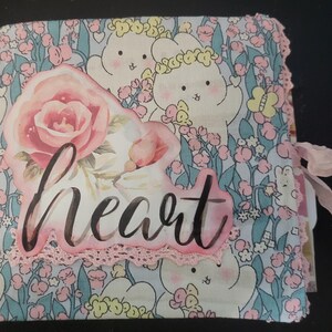 Pode incluir: Um álbum de recortes quadrado revestido de tecido com um fundo azul claro e um padrão repetitivo de coelhinhos brancos, flores cor-de-rosa e folhas verdes. Um recorte grande de rosa rosa com a palavra heart em letra preta está centrado.