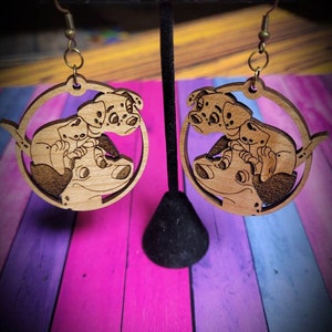 May include: A pair of wooden earrings featuring a cartoon image of Dalmatian puppies. The earrings are circular and have a brown wood finish.