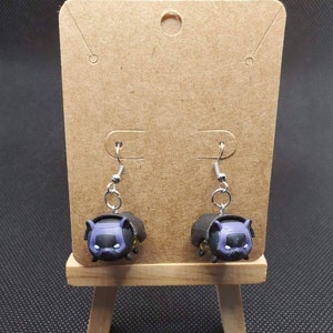 May include: A pair of dangle earrings featuring a black and purple cartoon cat with a yellow symbol on its chest. The cat is wearing a black cape and has a serious expression.