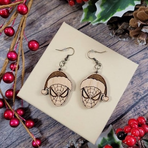 May include: A pair of wooden earrings featuring a Spiderman design with a Santa hat. The earrings are brown and have a gold hook.