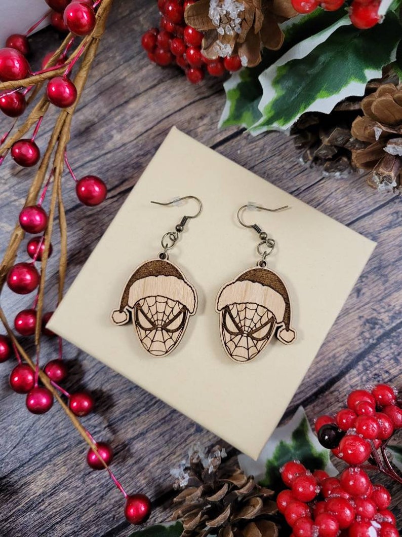 May include: A pair of wooden earrings featuring a Spiderman design with a Santa hat. The earrings are brown and have a gold hook.