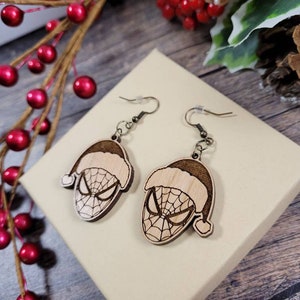 May include: A pair of wooden earrings featuring a Christmas-themed Spider-Man design. The earrings are brown and have a Santa hat on top of Spider-Man's head.