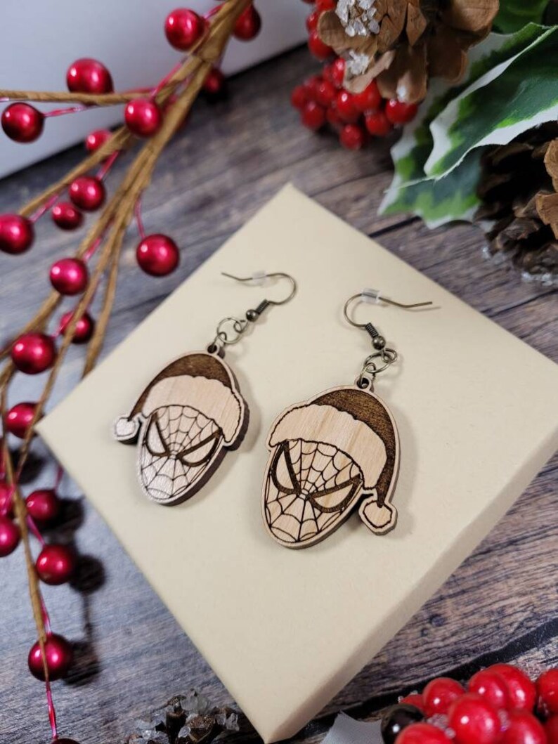 May include: A pair of wooden earrings featuring a Christmas-themed Spider-Man design. The earrings are brown and have a Santa hat on top of Spider-Man's head.