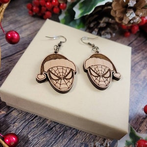 May include: A pair of wooden earrings featuring a Spiderman character wearing a Santa hat. The earrings are brown and have a detailed design.