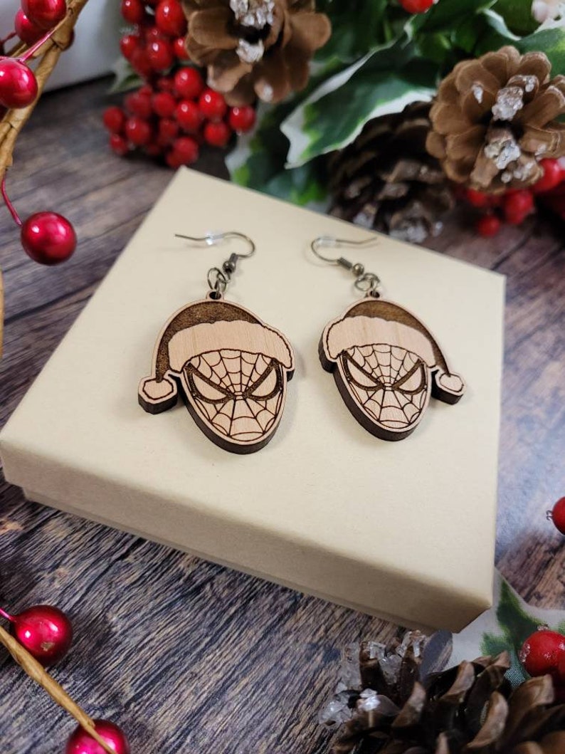 May include: A pair of wooden earrings featuring a Spiderman character wearing a Santa hat. The earrings are brown and have a detailed design.