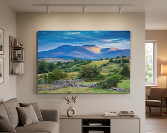 May include: A large rectangular landscape wall art print featuring a mountain range under a blue sky with golden sunlight hitting the peaks, above a green valley with rolling hills, scattered trees, and a low stone wall in the foreground.