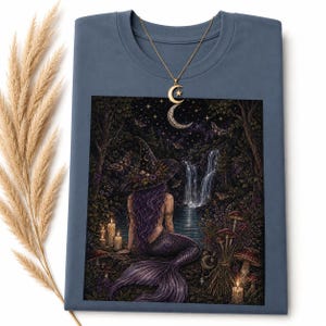 May include: A slate blue t-shirt featuring a graphic of a purple-haired mermaid wearing a witch hat, sitting by a waterfall at night with candles and mushrooms. A gold-toned necklace with a crescent moon and star pendant rests on the neckline.