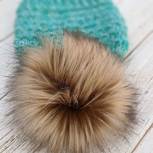 May include: A close-up of a teal and white knitted hat with a large, fluffy, light brown faux fur pom-pom. The pom-pom has dark brown tips. The hat is on a white wooden surface.