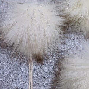 May include: Two white fluffy pom poms with a white string attached to one of them. The pom poms are on a gray textured background.
