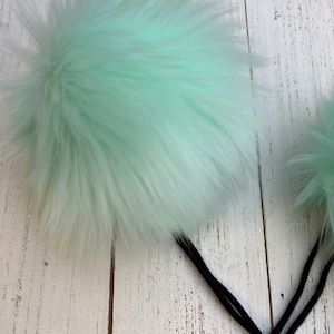 May include: Two fluffy mint green pom poms attached to black cords. The pom poms are soft and fuzzy, and the cords are long and thin.
