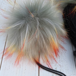 May include: A fluffy gray pom pom with red, orange, and yellow highlights. The pom pom is attached to a black cord.
