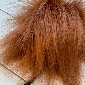 May include: A close-up of a fluffy, brown pom-pom with long, textured strands. The pom-pom is attached to a thin, black cord. The background is a white, wooden surface.
