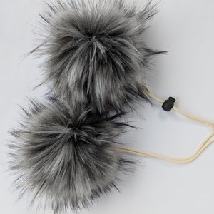 May include: Two black and white faux fur pom poms attached to a white cord with a black plastic toggle.