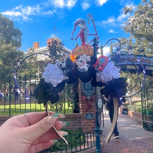 May include: Black and white Mickey Mouse ears headband with a black bow, white flower, and three Halloween-themed charms. The charms are a ghost, a heart, and a skeleton. The headband is in front of a Haunted Mansion entrance at Disneyland.