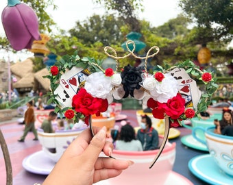 May include: A pair of black headband mouse ears with a floral design featuring red, white, and black roses, green leaves, and two playing cards with the Queen of Hearts and Jack of Hearts.