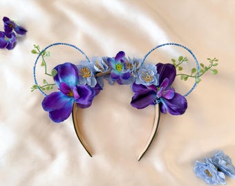 Peut inclure: Un serre-tête de fleurs violet et bleu avec un cadre en fil de fer. Le serre-tête est décoré de fleurs violettes et bleues et de feuilles vertes.