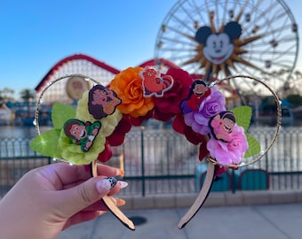 May include: A pair of gold Mickey Mouse ears with a floral headband featuring colorful flowers and character charms. The headband is decorated with red, orange, yellow, green, purple, and pink flowers. The charms are of cartoon characters with various expressions.