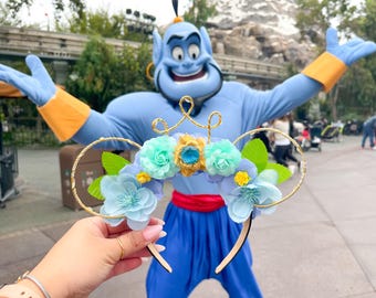 May include: A pair of gold Mickey Mouse ears with a blue and green floral design. The flowers are made of fabric and have a blue, green, and yellow color scheme. The ears are decorated with a gold crown and a blue gem.