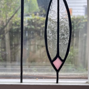 May include: A stained glass window panel featuring a vertical, elongated oval shape with textured, clear glass panes. The design includes a small, light pink diamond-shaped piece at the bottom and a vibrant green glass section at the top, all framed in black.
